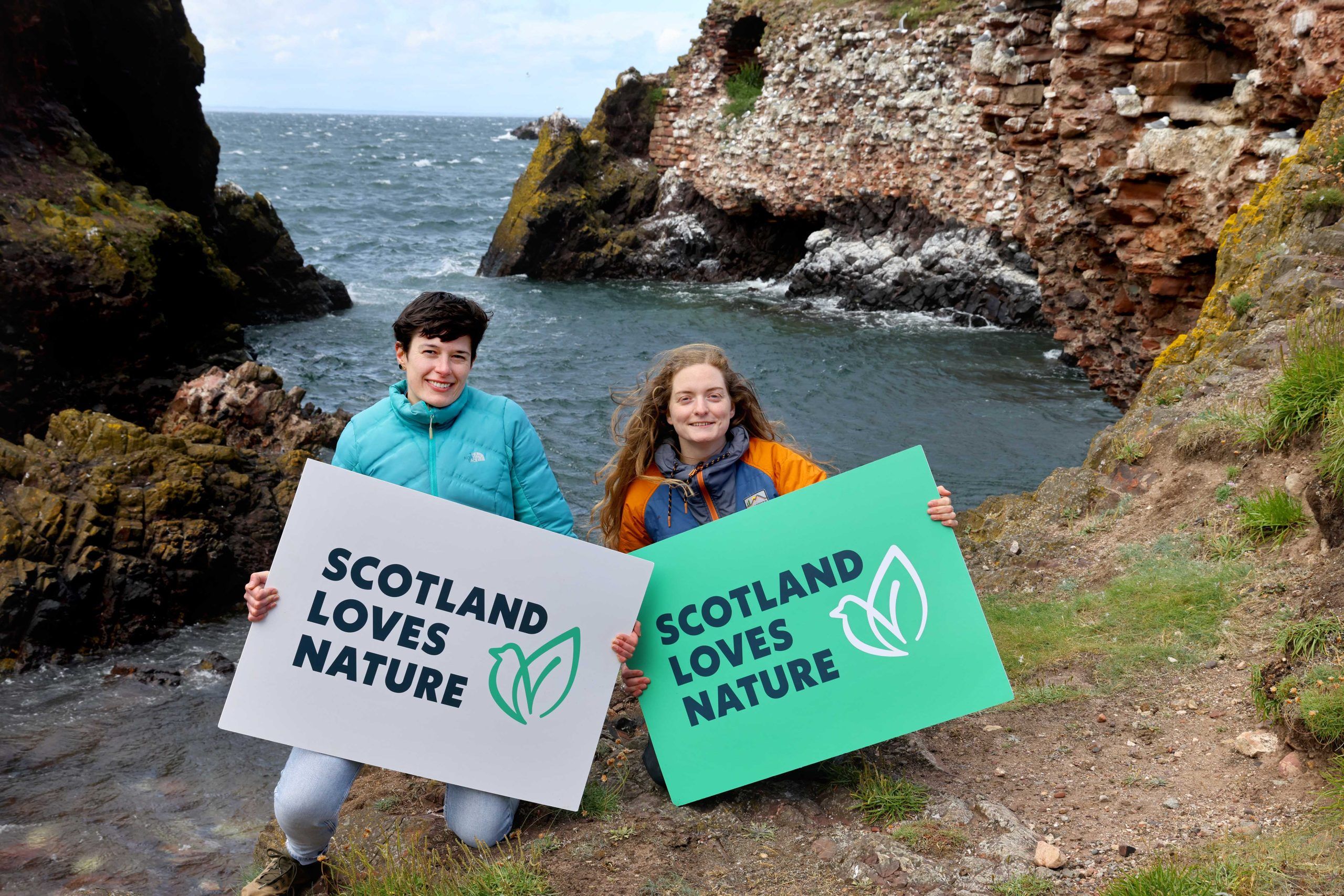 Two people holding signs which read "SCOTLAND LOVES NATURE" in front of a beach cove background